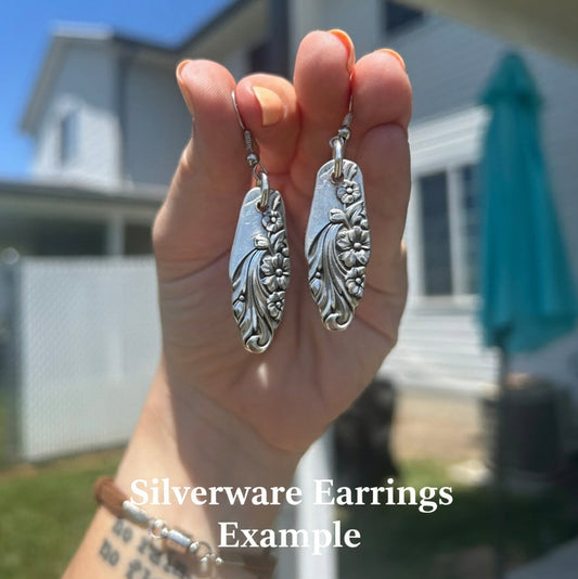 Hand holding two silver silverware earrings with a blurred outdoor background