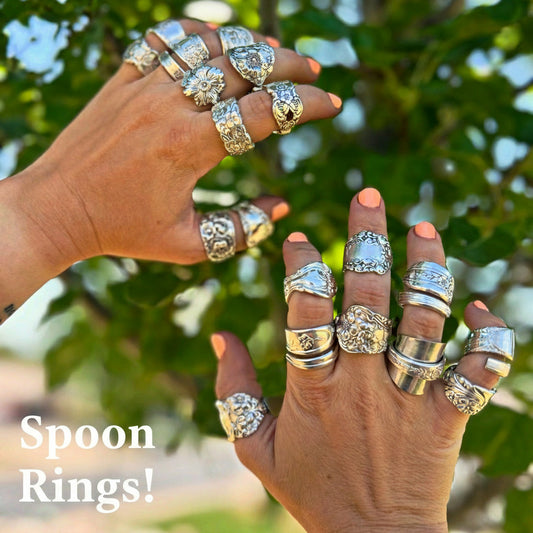 Two hands wearing multiple silver spoon rings with a blurred green leafy background