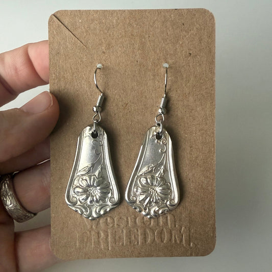Silver earrings on a brown card held by a hand with a ring.